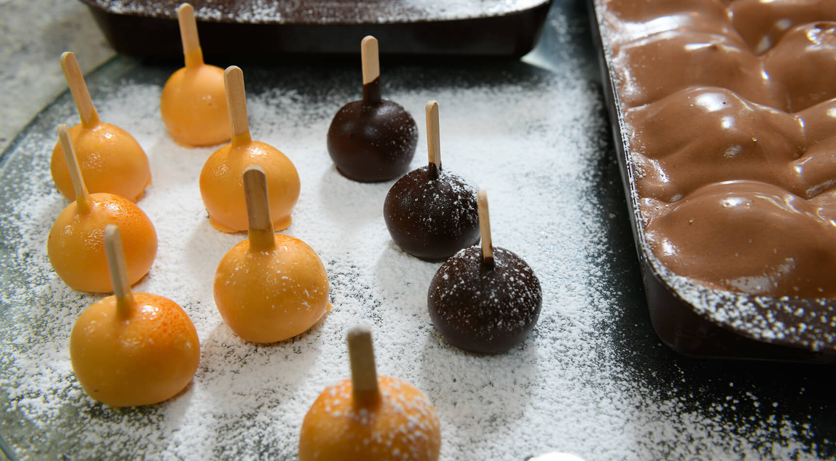 Orange und braune Kugeln auf Stäbchen, Puderzucker.