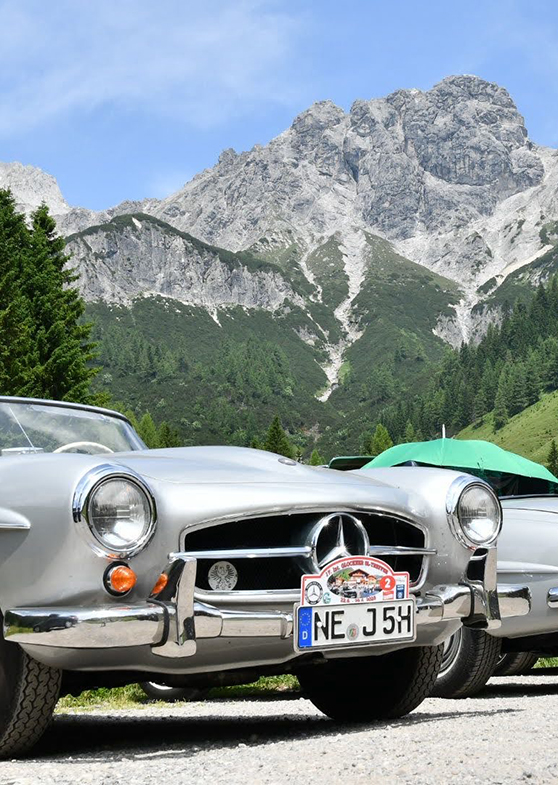 Zwei silberne Oldtimer vor grauen Bergen.