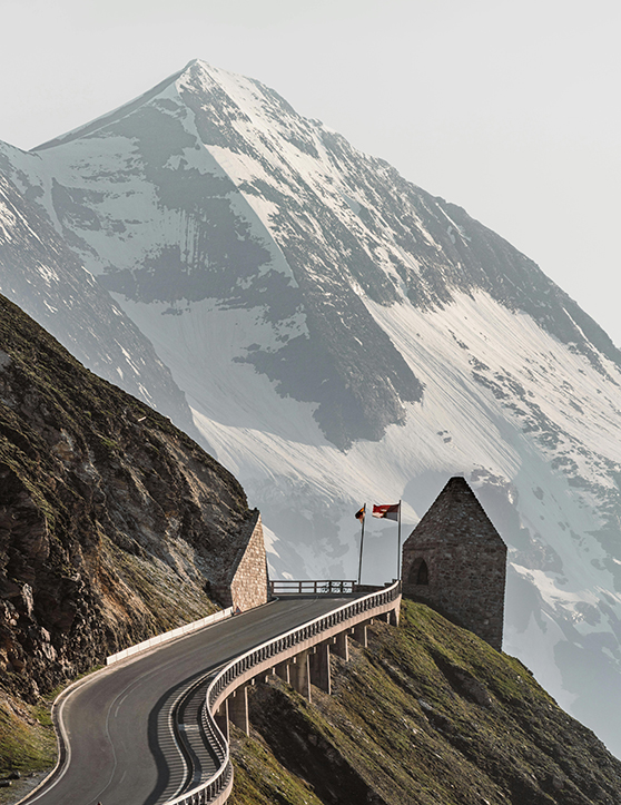 Bergstraße, Steinturm, zwei Flaggen, schneebedeckter Gipfel.