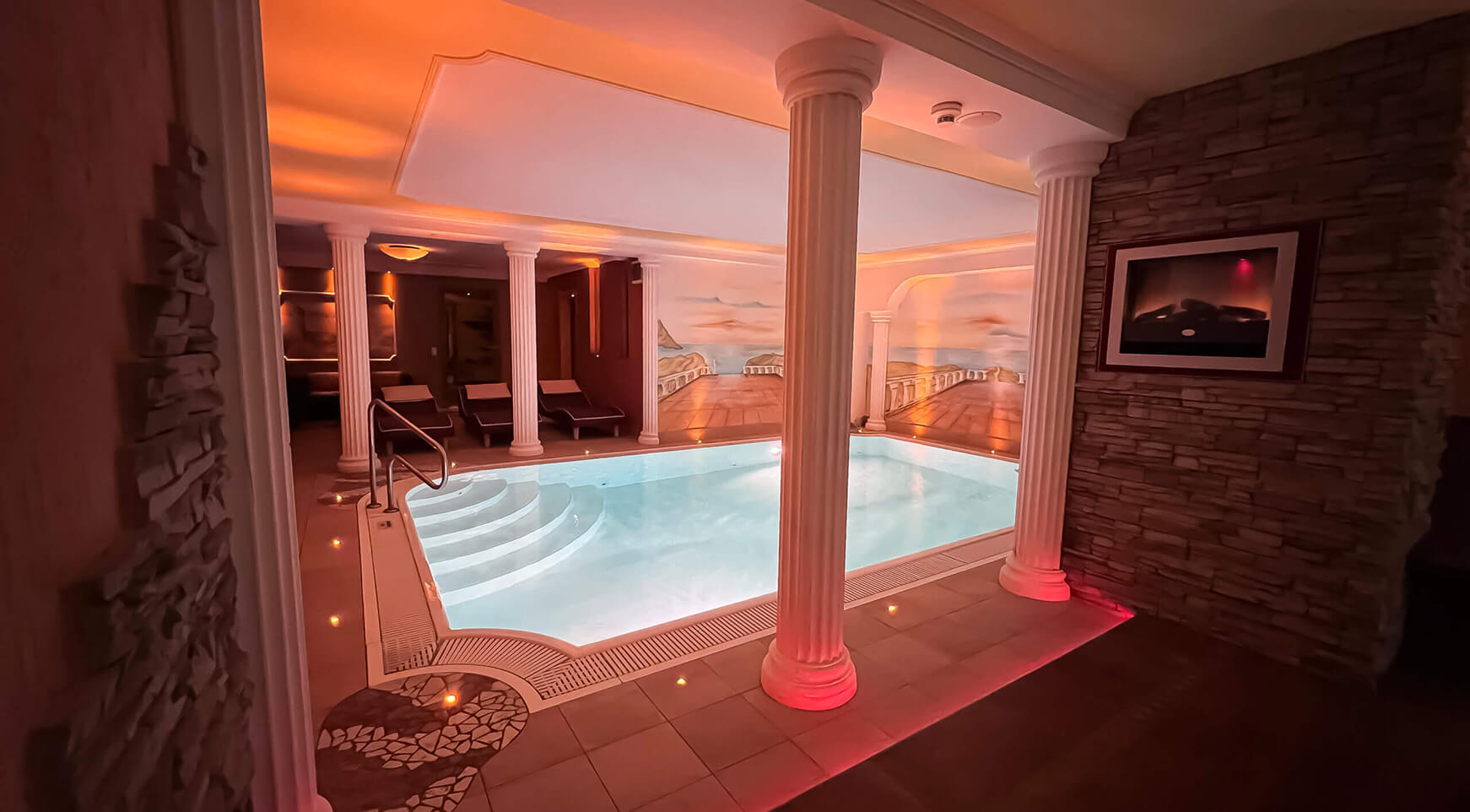 Innenpool mit Säulen, Wandbild und warmem Licht.