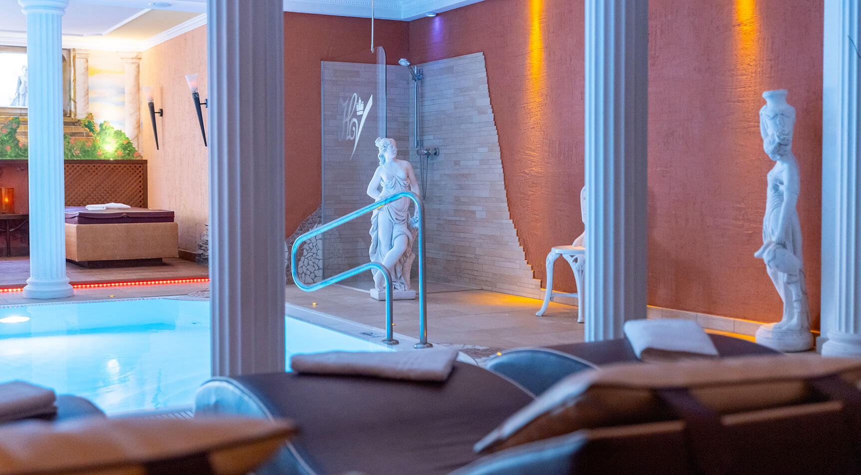 Innenpoolbereich mit Säulen, Statuen und Liegen.